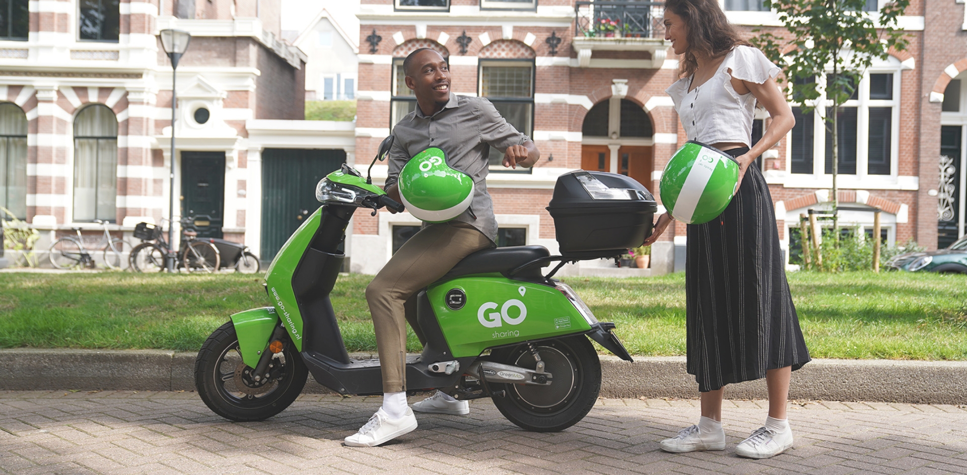A man sitting on a parked GO Sharing scooter. He is looking back to a woman standing behind the scooter. Both are holding a GO Sharing helmet.